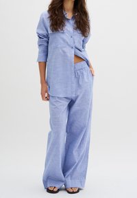 Světle modrá, pruhovaná košile s dlouhými rukávy a kapsou, doplněná volnými kalhotami ve stejném odstínu a plochými černými sandály.