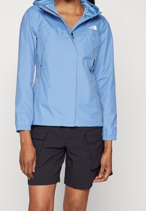 Vrouw draagt een lichtblauw regenjack met capuchon, rits en stormklep, gecombineerd met zwarte cargoshorts tegen een effen achtergrond.