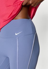 Nike legíny s vysokým pasem v světle modré barvě s kontrastním bílým logem. Hladký povrch a postranní švy pro zvýraznění detailů. Viditelný růžový top.
