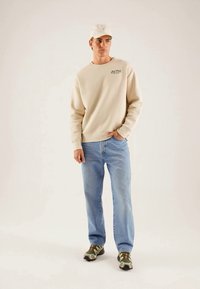 Kremfarget sweatshirt med rund hals, kombinert med lyseblå bukser. Modellen har på seg en hvit caps og grønne camo-sneakers, med hendene i lommene.