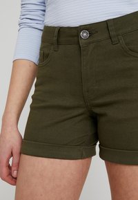 Pantalones cortos de mezclilla verde oliva con dobladillos enrollados, cierre frontal con botón y bolsillos laterales. Textura suave y diseño ajustado.