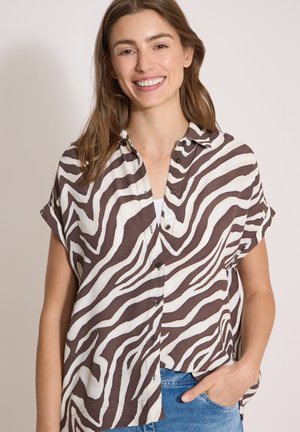 Junge Frau lächelt, trägt ein kurzärmeliges Hemd mit schwarz-weißen Zebrasteifen und blaue Jeans, Hand in der Tasche.
