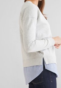 Sweat-shirt gris clair avec un col rond, des poignets côtelés et des fentes latérales, porté sur une chemise bleu clair à col avec un léger motif.