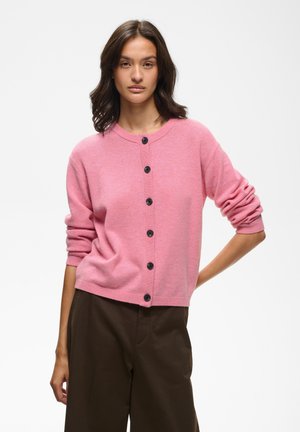Jeune femme portant un cardigan rose boutonné avec les manches remontées et un pantalon marron foncé sur un fond blanc uni.