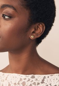 Goldener Ohrstecker in einzigartiger Herzform mit polierter Oberfläche, getragen von einem Modell mit kurzem, strukturiertem Haar, vor neutralem Hintergrund.