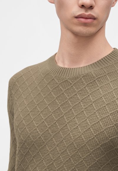 Jeune homme portant un pull en maille texturée marron clair avec un motif en losange et un col rond côtelé.
