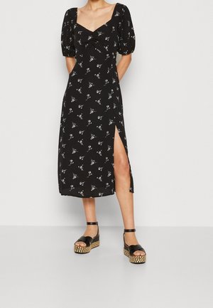 Sort blomsterkjole med korte puffede ærmer, firkantet udskæring, knudedetalje ved busten og sideslids; kombineret med sorte platformsandaler.