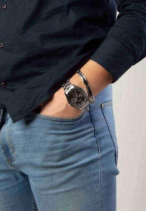 Silver stainless steel watch with a black dial, chronograph features, and a textured metal bracelet. Worn with blue denim jeans.