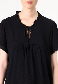 Blusa nera a maniche corte con colletto arricciato, apertura a chiave legata con un fiocco e tessuto testurizzato con piccole palline imbottite.