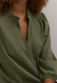 Personne aux cheveux bouclés portant un chemisier côtelé vert olive avec un col à volants et des manches bouffantes sur un fond clair uni.