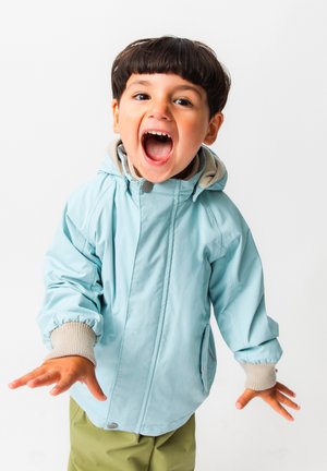 Noor laps lühikeste tumedate juustega, kandes helesinist jopet ja rohelisi pükse, suu lahti, käed sirutatud ettepoole.