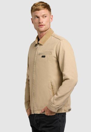 Muž v béžovej bunde na zips s manžetou z kordurovej látky a čiernymi nohavicami, stojaci s rukami v vreckách, pozerajúci sa do strany.