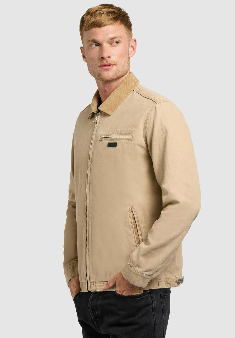 Homme portant une veste zippée beige avec un col en velours côtelé et un pantalon noir, debout les mains dans les poches, regardant sur le côté.