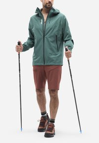 Veste imperméable verte avec une capuche, deux poches latérales et une fermeture éclair ; associée à un short couleur rouille et des chaussures de randonnée marron, utilisant des bâtons de trekking.