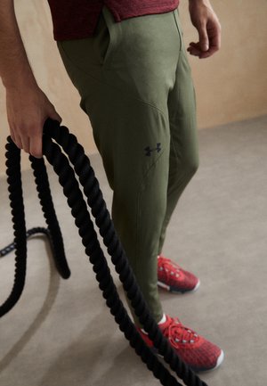 Person trägt olivgrüne Under Armour Hose und rote Sneaker und hält gekringelte schwarze Battle Ropes in einem Trainingsumfeld.