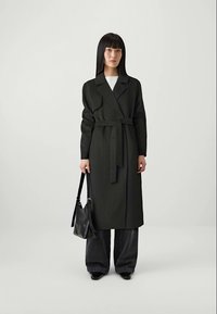 Donna che indossa un lungo cappotto nero con cintura, jeans neri a gamba larga, scarpe nere, e tiene in mano una borsa nera su uno sfondo chiaro e semplice.
