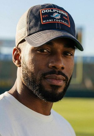 Man with beard wearing a Miami Dolphins baseball cap and white shirt, outdoors on a sunny day with blurred background.