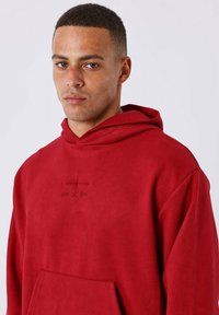 Sudadera roja fabricada con un tejido suave, con un bolsillo canguro, presenta un logotipo bordado en el centro y una capucha grande y holgada.