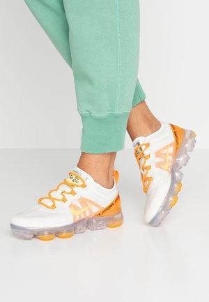 Stopy w białych i pomarańczowych sneakersach z przezroczystą podeszwą, w zielonych spodniach dresowych, stojące na gładkiej, refleksyjnej podłodze.