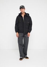 Black hooded jacket with multiple front pockets, paired with gray pants and black sneakers featuring white accents, standing on a plain background.