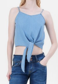 Crop top en denim bleu clair avec bretelles spaghetti ajustables et détail noué à l'avant. Porté avec un jean en denim bleu, mettant en valeur un style décontracté.
