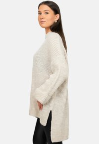 Licht beige gebreid sweater met een losse pasvorm, geribde manchetten, zijsplitten en een gestructureerd patroon. Draagt over zwarte legging.