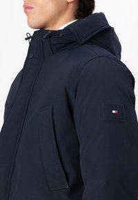 Giacca a cappuccio blu navy realizzata in tessuto liscio; presenta una tasca laterale, una patta e una toppa con logo sulla manica.