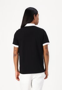 Μαύρο πουκάμισο polo με κοντά λευκά κολάρα, από βαμβακερό υλικό, με χαλαρή εφαρμογή, απεικονίζοντας την πίσω πλευρά και τον μινιμαλιστικό σχεδιασμό.