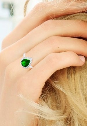 Emerald green pear-shaped gemstone ring with a silver band, surrounded by small clear stones, held against a light skin tone.