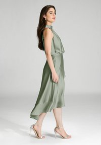 Robe en satin vert avec un col haut, sans manches, jupe fluide et fente haute. Associée à des sandales à talons hauts argentées.
