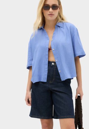 Kvinde iført mørke knælange denimshorts, løs lyseblå kortærmet skjorte med knapper, solbriller og holder en mørk flettet taske.