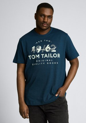 Mężczyzna stojący z ręką w kieszeni, ubrany w granatowy t-shirt Tom Tailor z napisem "1962" i tekstem "Original Quality Goods" na przodzie, szare tło.