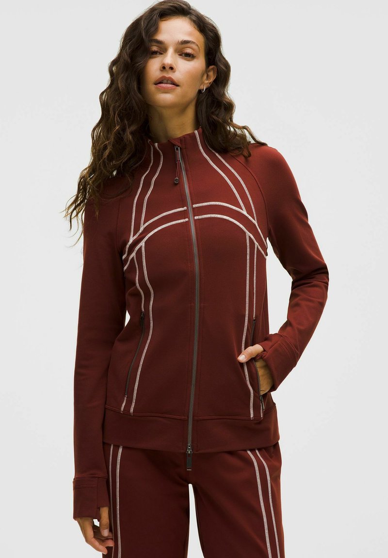 Felpa rossa con zip realizzata in un tessuto liscio e morbido, caratterizzata da linee curve bianche e tasche laterali. Maniche lunghe con fori per il pollice per una maggiore copertura.