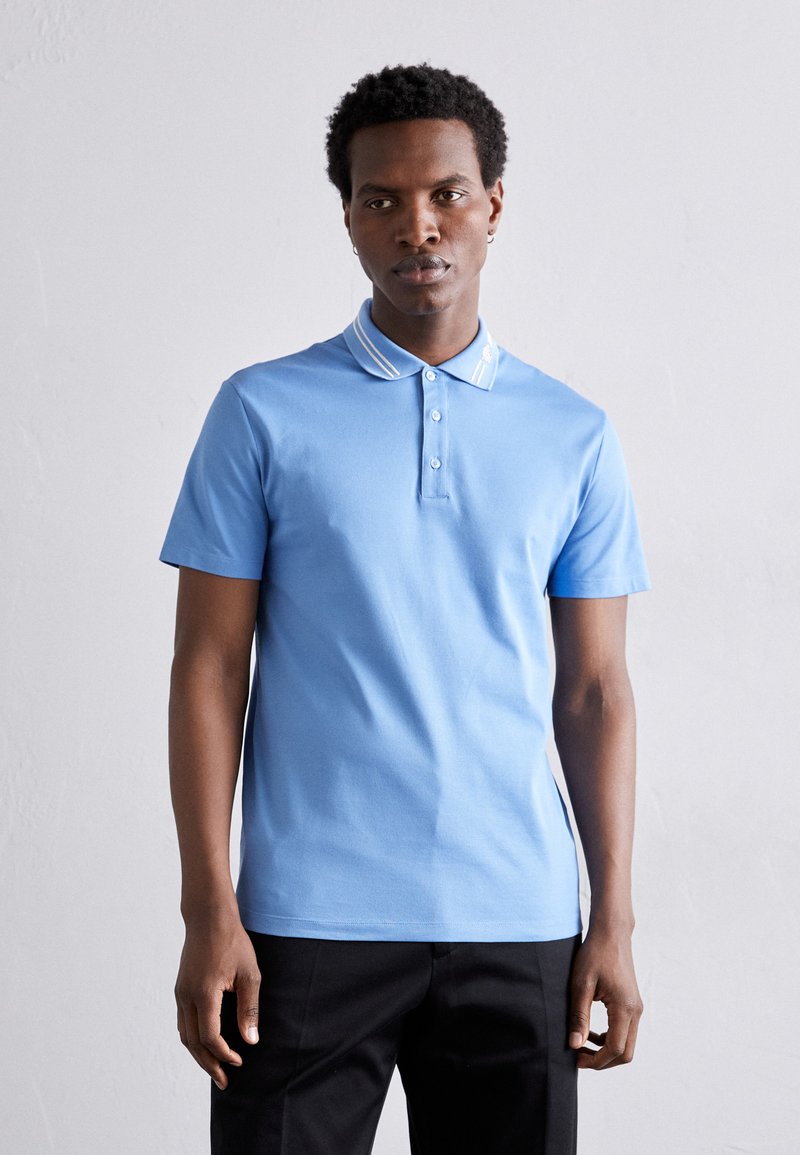 Polo de manga curta azul claro com pala de dois botões e colarinho listrado branco em contraste. O material parece suave e respirável.