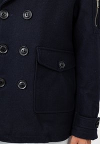 Navy niebieski wełniany płaszcz z podwójnym zapięciem, dużymi czarnymi guzikami, kieszenią z klapką oraz szczegółami na mankietach. Miękka, fakturowana powierzchnia.
