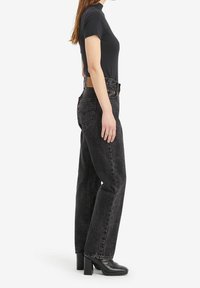 Mujer con un top negro de manga corta y cuello alto, jeans negros de cintura alta desgastados y botines negros de tacón, de pie de lado sobre fondo blanco.