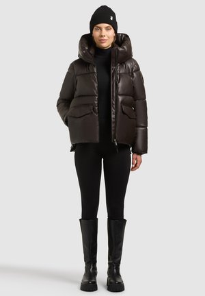 Femme portant un bonnet noir, une doudoune marron foncé, des leggings noirs et des bottes noires montantes, debout sur un fond gris clair.