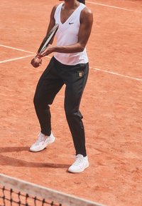 Bílé sportovní tílko bez rukávů, černé sportovní kalhoty s logem a bílé tenisky s růžovými akcenty na hlinitém tenisovém kurtu.