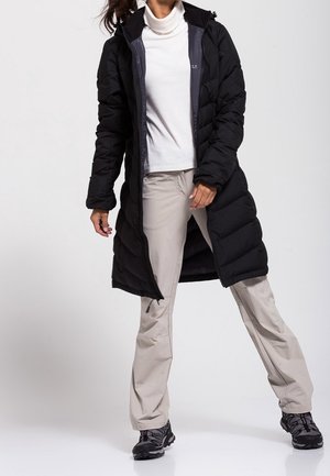 Femme debout portant un long manteau matelassé noir sur un col roulé blanc, un pantalon beige et des chaussures de randonnée noires, sur un fond uni.