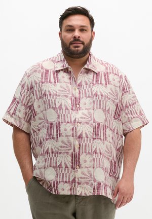 Brodaty mężczyzna w luźnej, krótkim rękawie koszuli zapinanej na guziki z różowo-białym kwiatowym i paskowanym wzorem, ręce częściowo w kieszeniach.