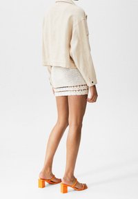 Cropped beige denim jacket, white crocheted mini skirt, and orange block-heeled sandals with slim straps. Textured and layered look.
