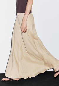 Maxi skirt in patterned beige fabric, featuring a flowing design, complemented by dark brown flat sandals. Elegant texture and silhouette.