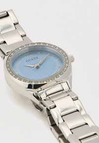 Sølvfarvet rustfrit stålkasse ur med rund blå urskive, glitrende krans og forhøjede tal. Har et sammenkoblet metalarmbånd.