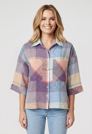 Femme souriante aux cheveux blonds portant une chemise ample, boutonnée, à carreaux multicolores et un jean bleu clair, debout devant un fond uni.