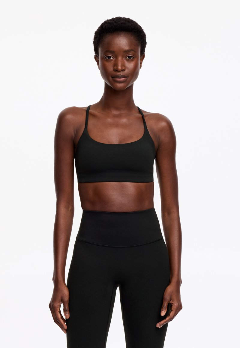 Femme aux cheveux courts et bouclés portant un soutien-gorge de sport noir et un legging noir taille haute, debout sur un fond blanc.