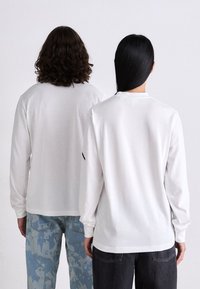 Cămăși albe cu mâneci lungi, croi lejer. O persoană în blugi deschiși la culoare, cealaltă în blugi închiși. Design simplu, fără ornamentații, cu guler rotund.