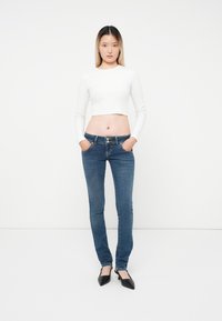 Fehér, cropped, hosszú ujjú felső, kék, testhez simuló farmernadrág, gombos záródással és két elülső zsebbel, fekete, hegyes orrú lapos cipőkkel párosítva.