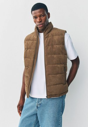 Brown padded vest with a high collar, soft texture, front zipper, and side pockets, worn over a white t-shirt and denim jeans.