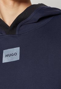 Sweat à capuche bleu marine avec un col côtelé noir et une étiquette rectangulaire bleu clair affichant "HUGO" en texte bleu foncé. Texture douce.