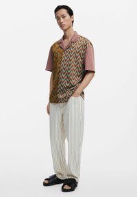 Short-sleeve shirt featuring a multi-patterned design in earthy tones, paired with white striped trousers and black sandals.